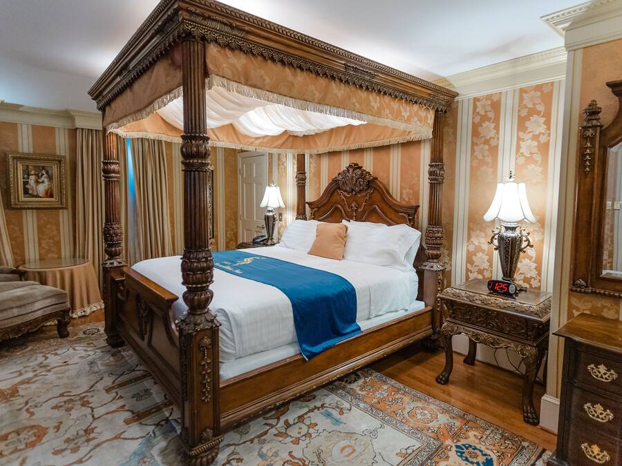 A four-poster bed with a canopy, a nightstand with a clock, a chair, and a mirror.