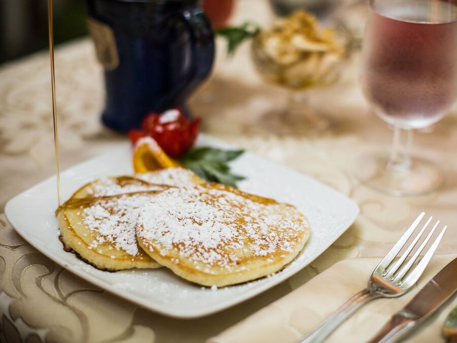 Een bord pannenkoeken met siroop en poedersuiker, een vork en mes op een tafel met een geruit tafelkleed.