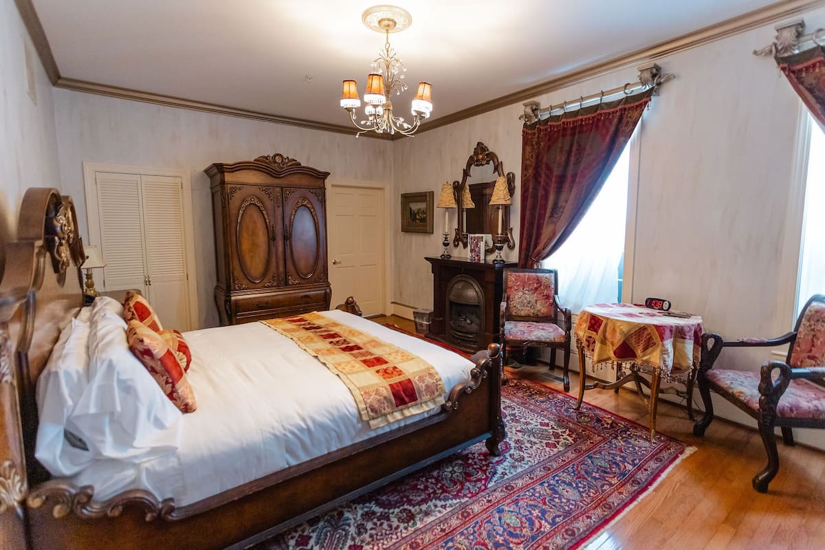 A bedroom with a large bed, a wooden wardrobe, a chandelier, a fireplace, and a small table with a clock.