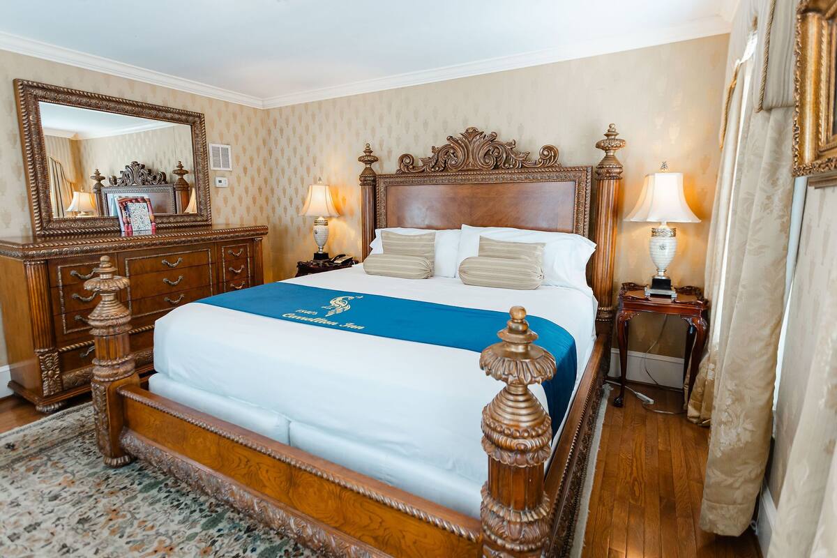 A large, ornate wooden bed with a blue bedspread, flanked by two nightstands with lamps, a mirror above the bed, and a wooden dresser with drawers.