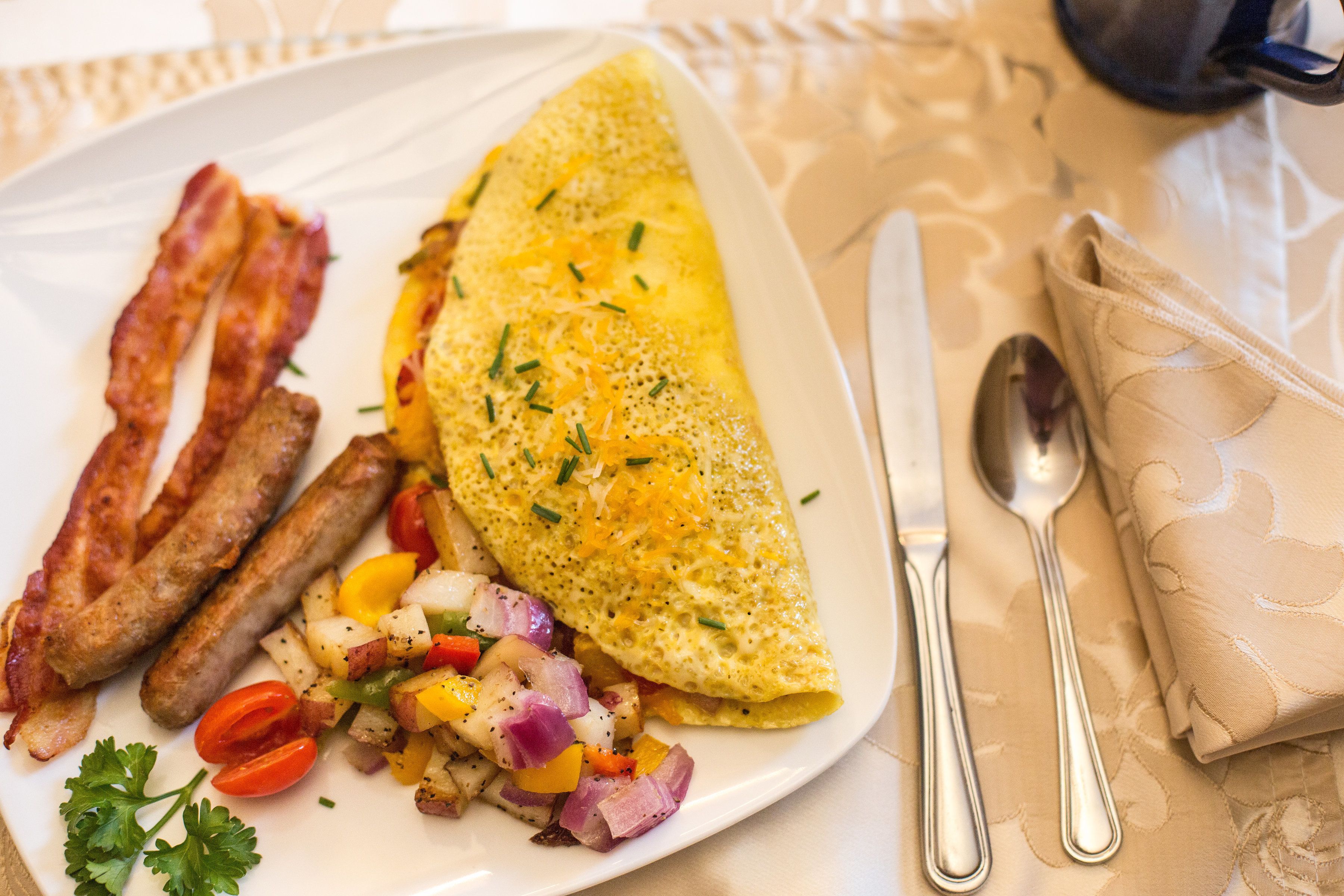A plate with an omelette, bacon, sausages, and diced vegetables.