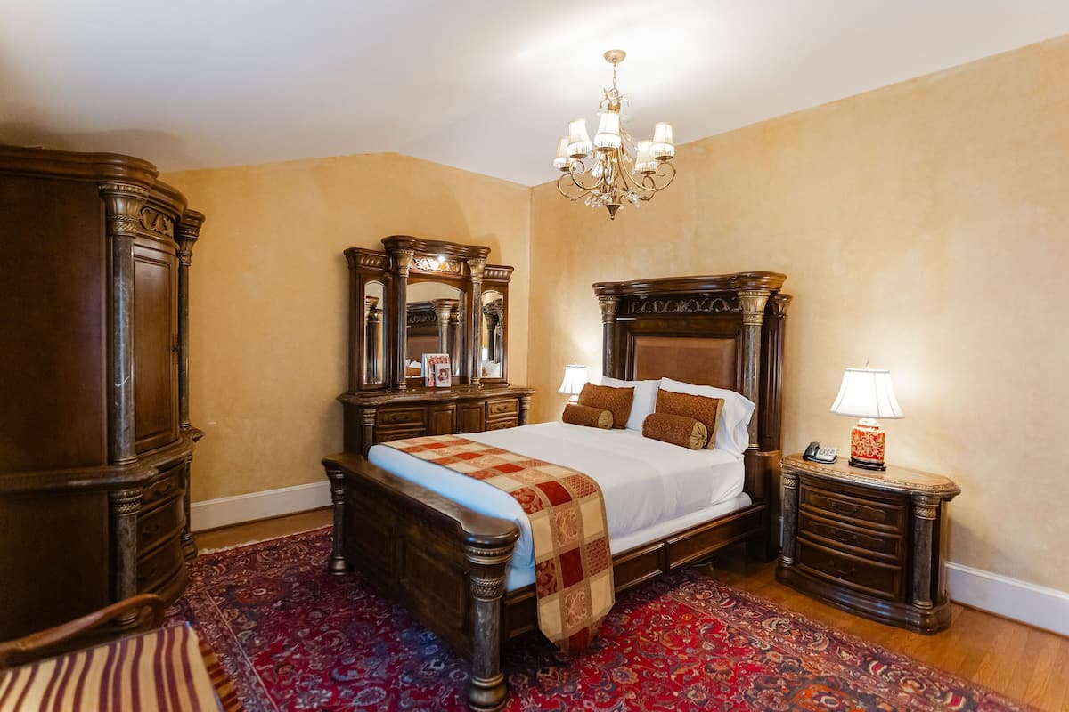 A traditional bedroom with a large bed, ornate wooden headboard, a chandelier, and a red patterned rug.