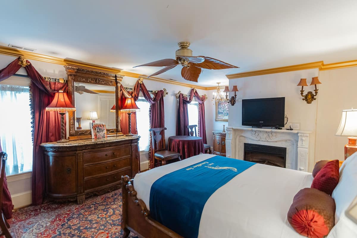A hotel room with a large bed, a wooden dresser, a television, a fireplace, and a ceiling fan.