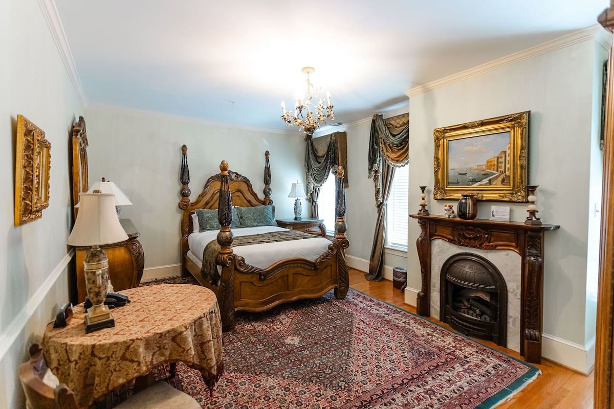 A bedroom with a four-poster bed, a chandelier, a fireplace, and a large rug.