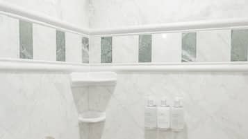 Casa de banho com chuveiro, azulejos de mármore branco e uma prateleira com artigos de higiene pessoal.