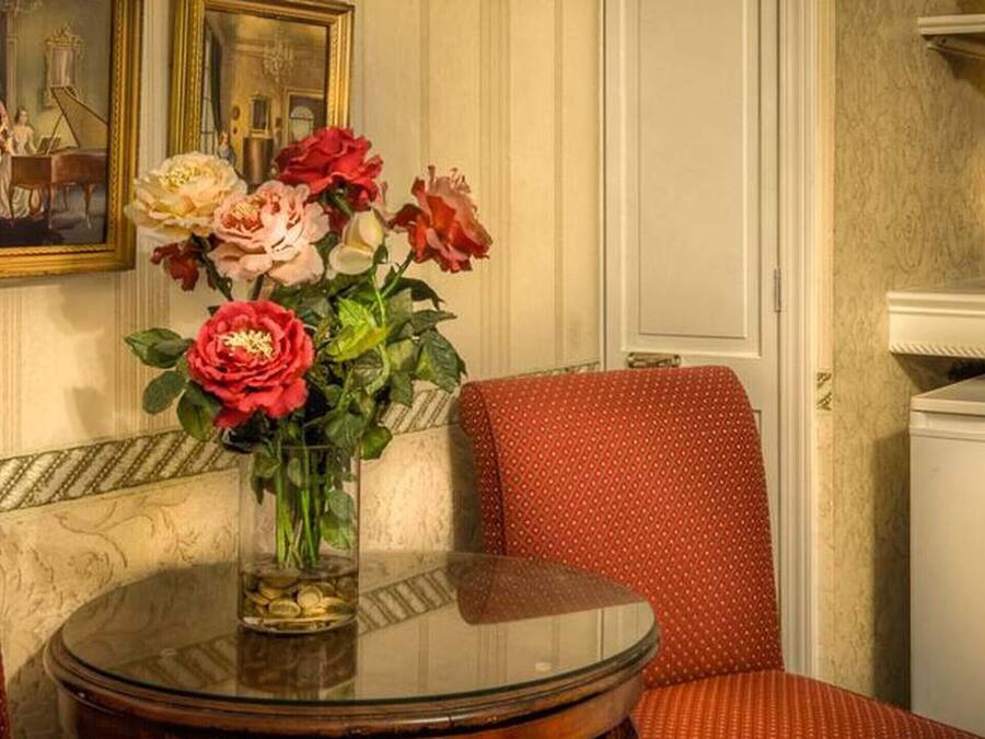 A room with a round glass table, red upholstered chairs, a vase with flowers, a painting on the wall, and a coffee machine.