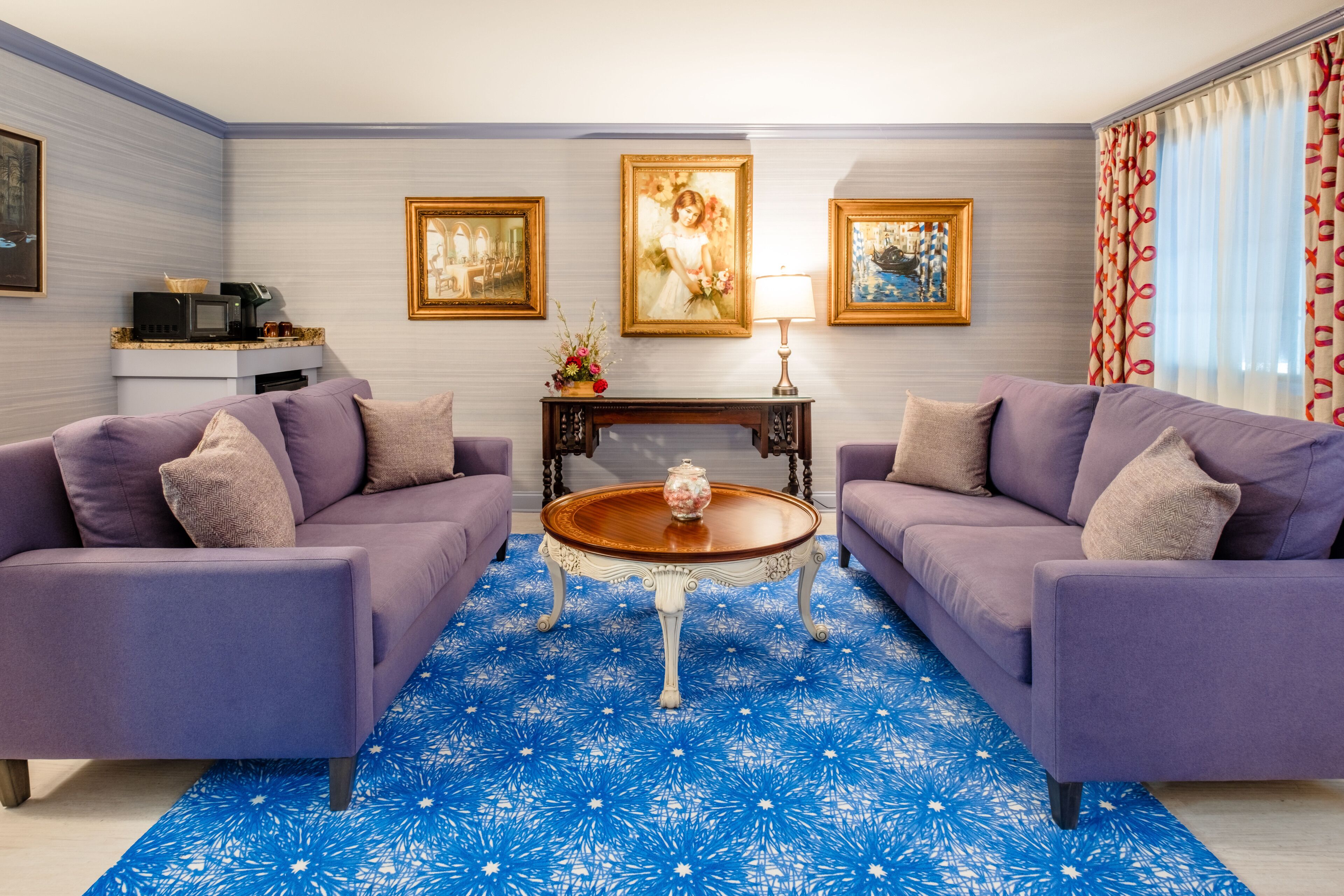 Een woonkamer met twee paarse banken, een houten salontafel en een blauw tapijt met een patroon. Er hangen ingelijste foto's aan de muur en staat een bijzettafeltje met een lamp en bloemen.