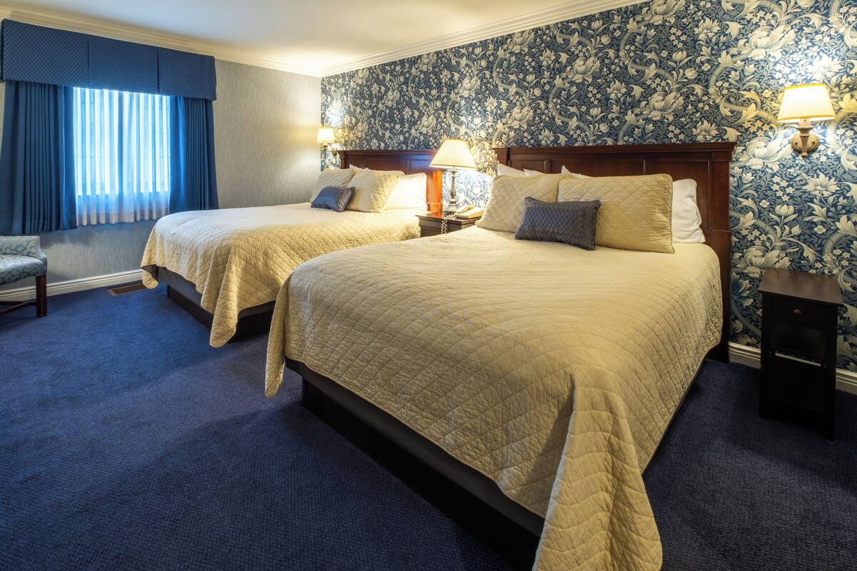 A hotel room with two beds, a window with blue curtains, a floral wallpaper, and a nightstand with a lamp.