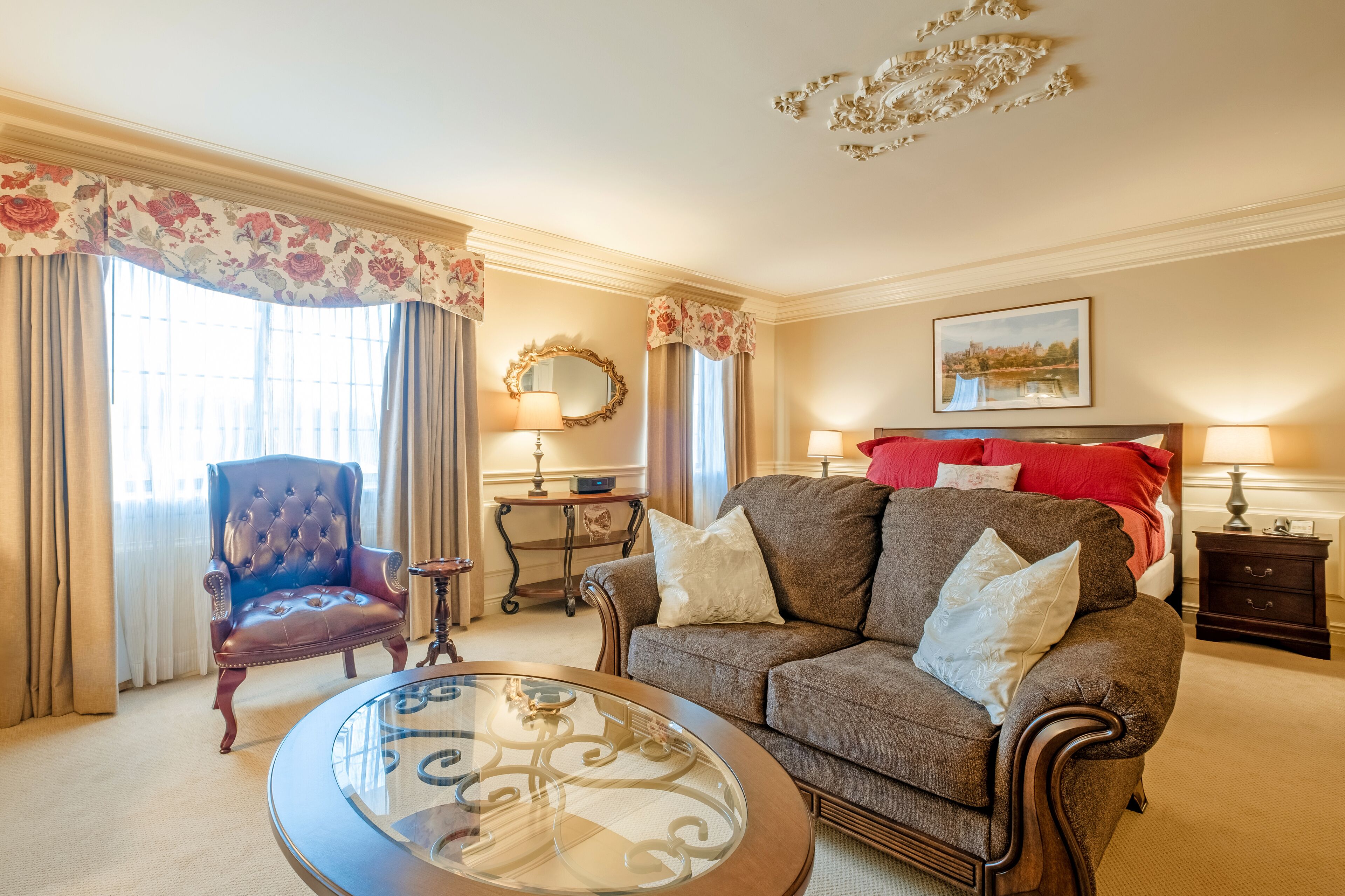A well-appointed hotel room with a tufted armchair, a sofa with white cushions, a round glass coffee table, and a bed with red pillows.