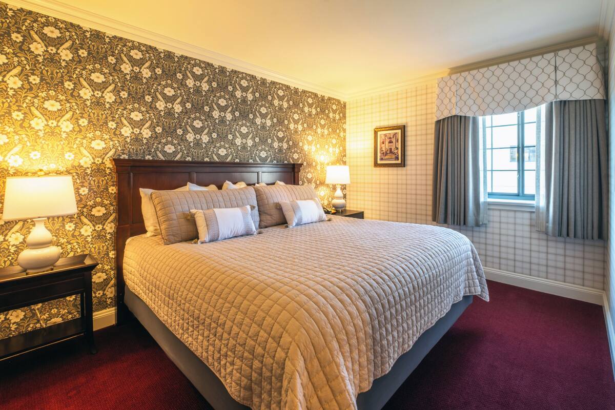 A neatly made bed with a quilted cover, two bedside tables with lamps, floral wallpaper, a framed picture on the wall, and a window with curtains.