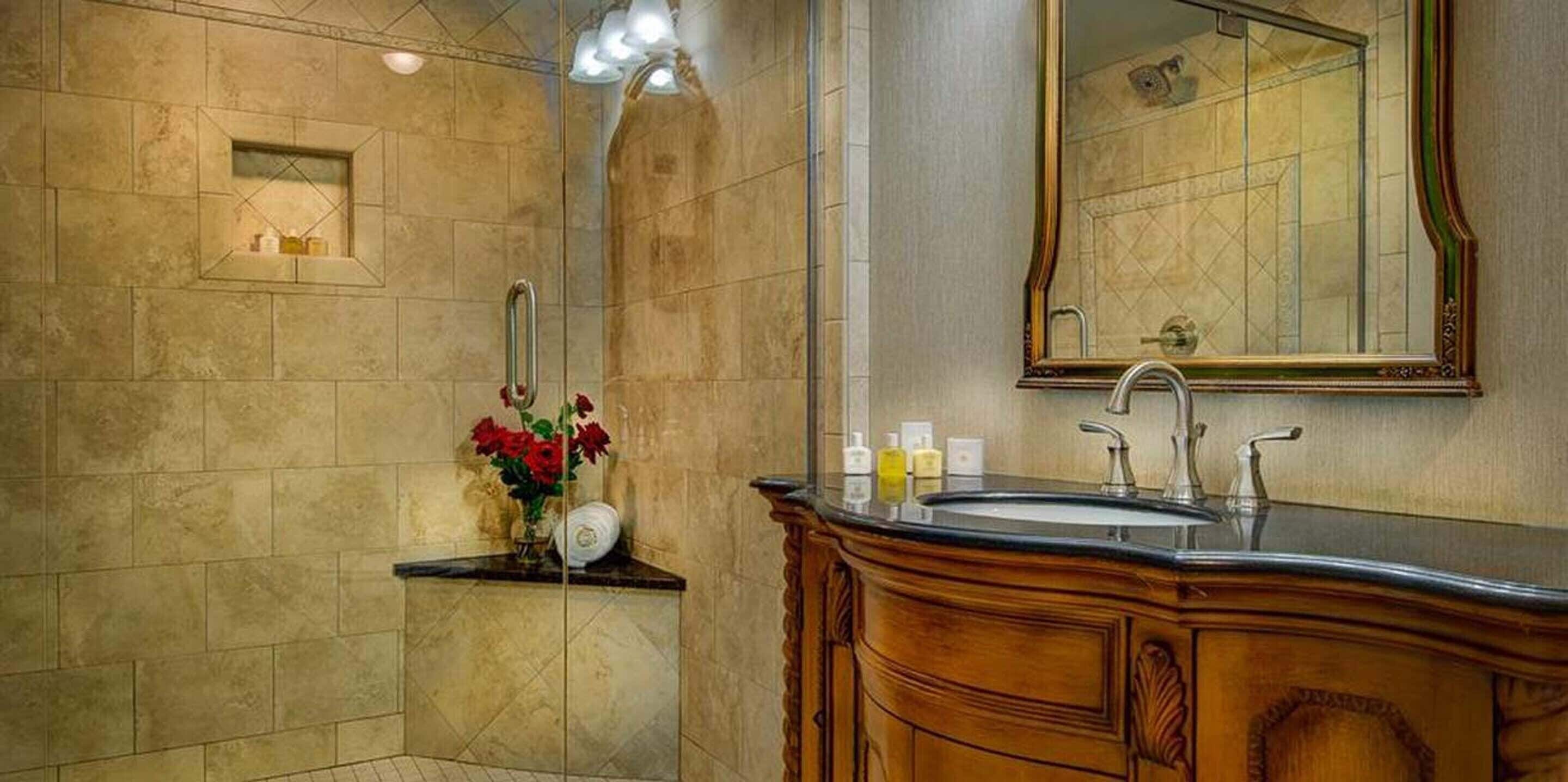 A bathroom with a large mirror, a sink, and a vanity with a mirror. There is a small shelf with toiletries and a vase with flowers.