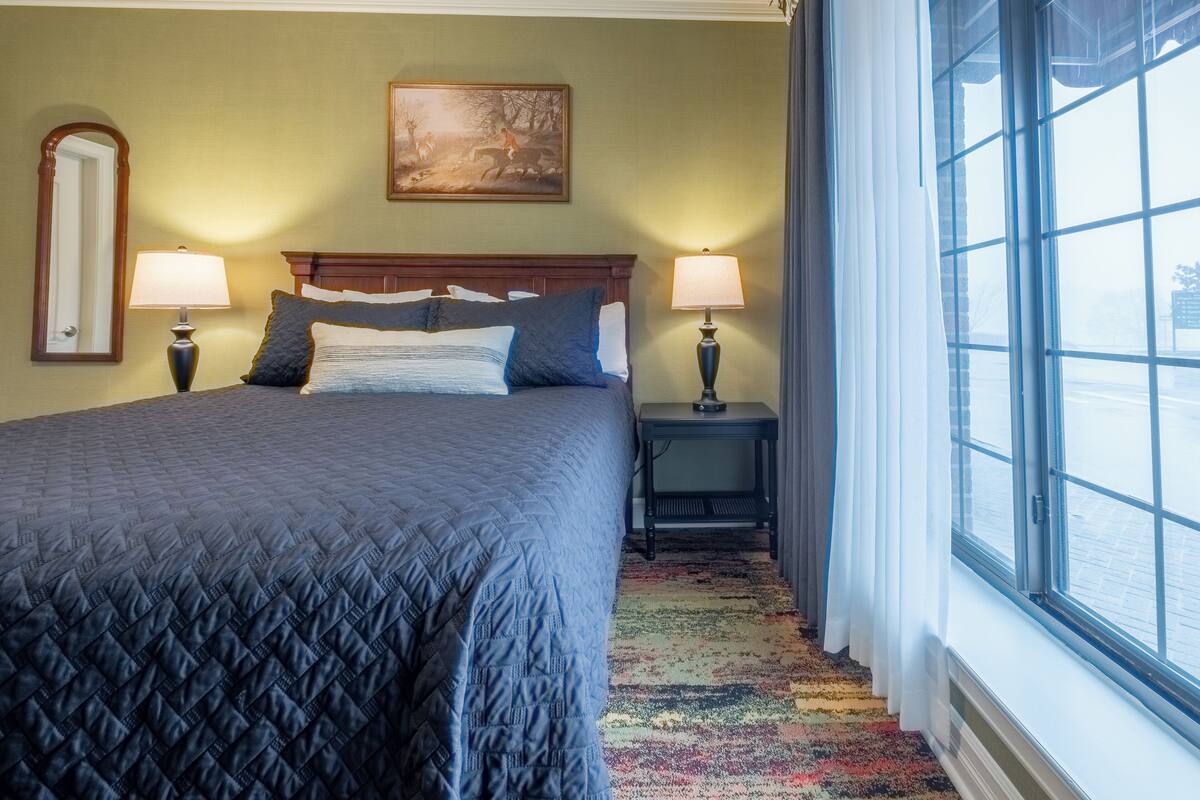 A hotel room with a large bed, two bedside lamps, a painting on the wall, and a window with curtains.