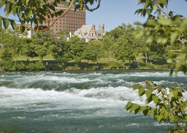 A river with white water rapids, surrounded by lush green trees and a historic building with a tower in the background. - Red Coach Inn (Niagara Falls)