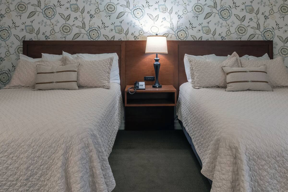 A hotel room with two beds, a nightstand with a lamp, and floral wallpaper.