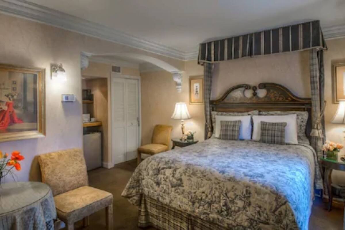 A bedroom with a four-poster bed, a chair, a table with flowers, and a painting on the wall.