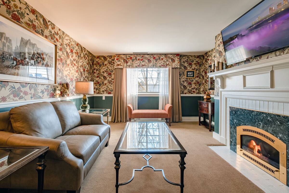 A living room with a leather sofa, a glass-top coffee table, a fireplace, and floral wallpaper.