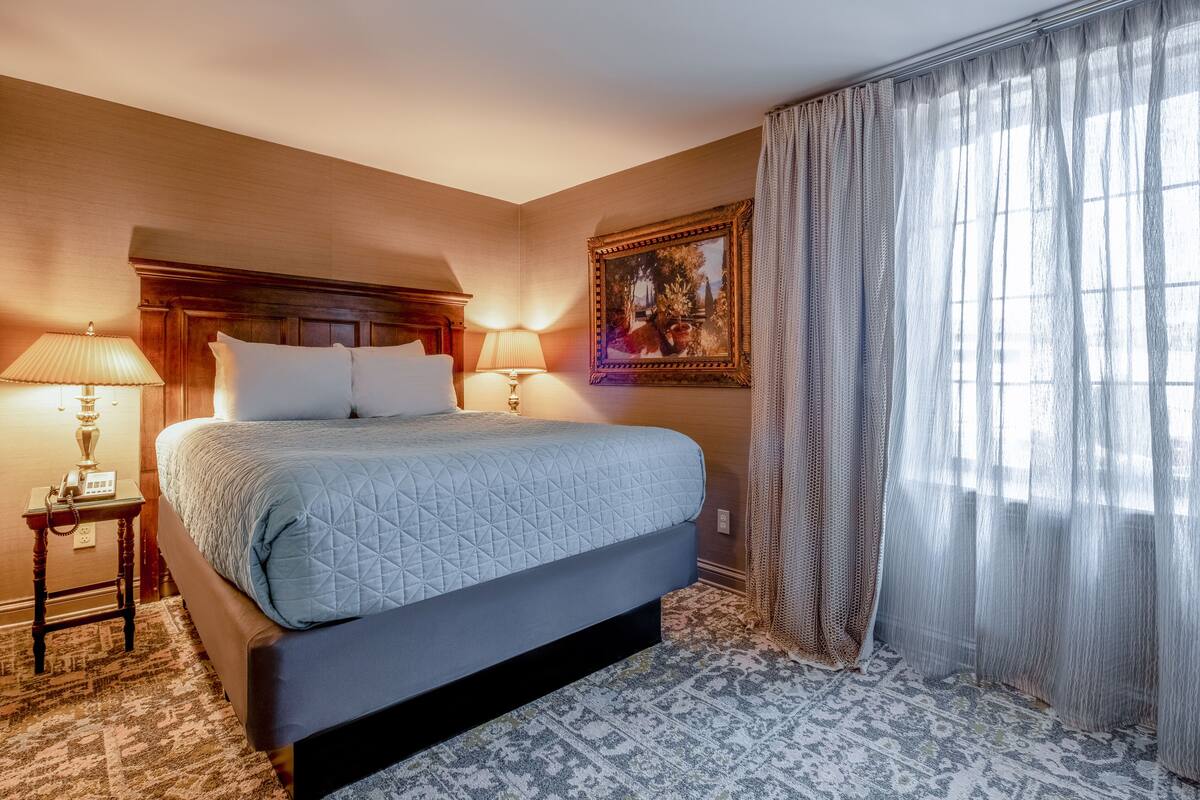 A hotel room with a large bed, two bedside lamps, a painting on the wall, and sheer curtains.