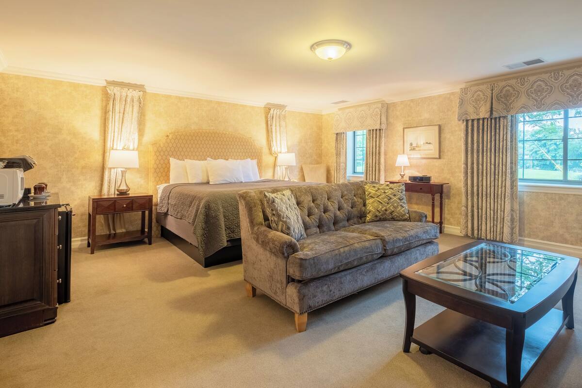 A hotel room with a large bed, a sofa, a glass-top coffee table, and a small side table with a lamp.