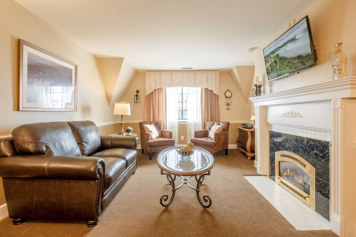 A living room with a fireplace, leather sofas, a glass coffee table, and a television mounted above the fireplace.