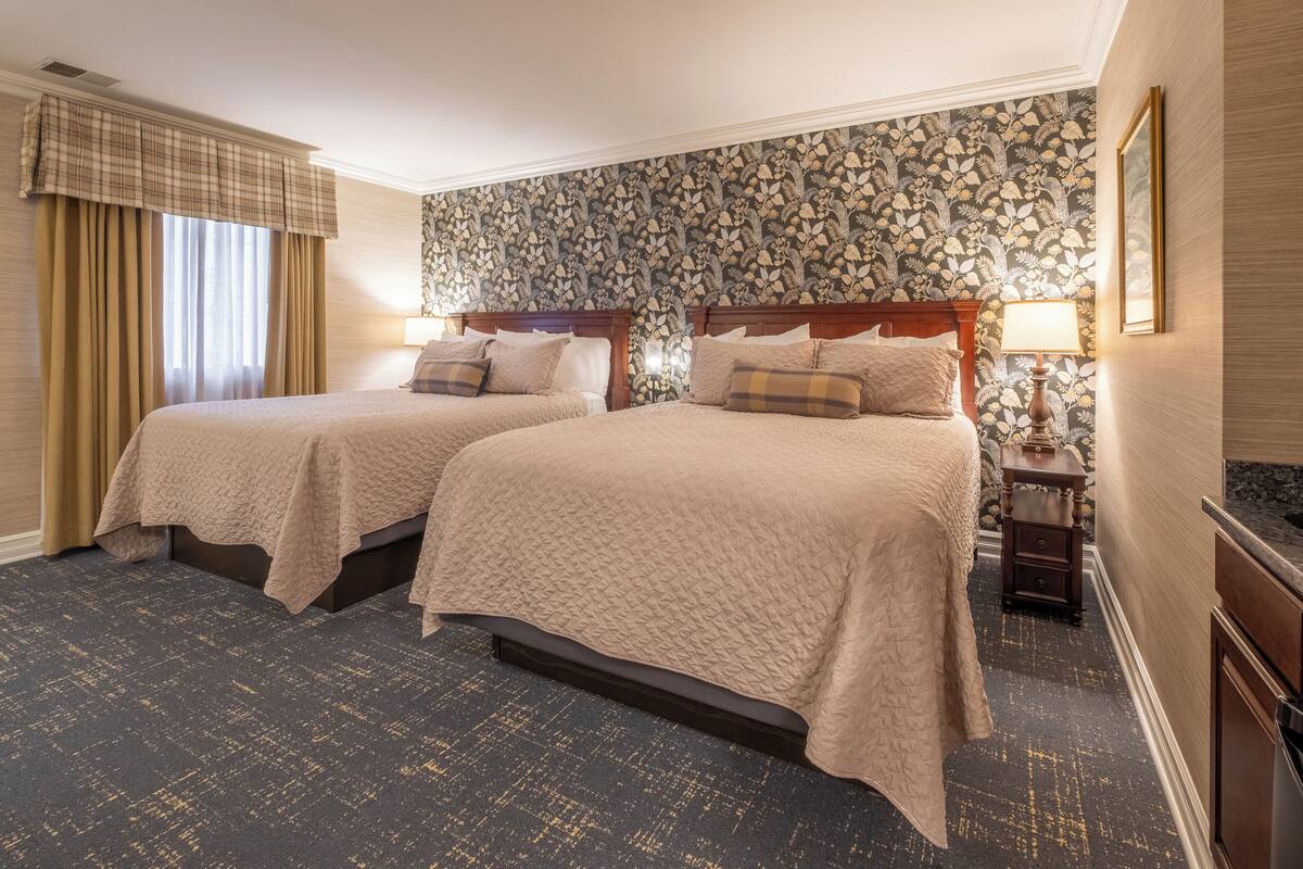 A hotel room with two beds, a large window with curtains, a floral wallpaper, and a dark carpet.