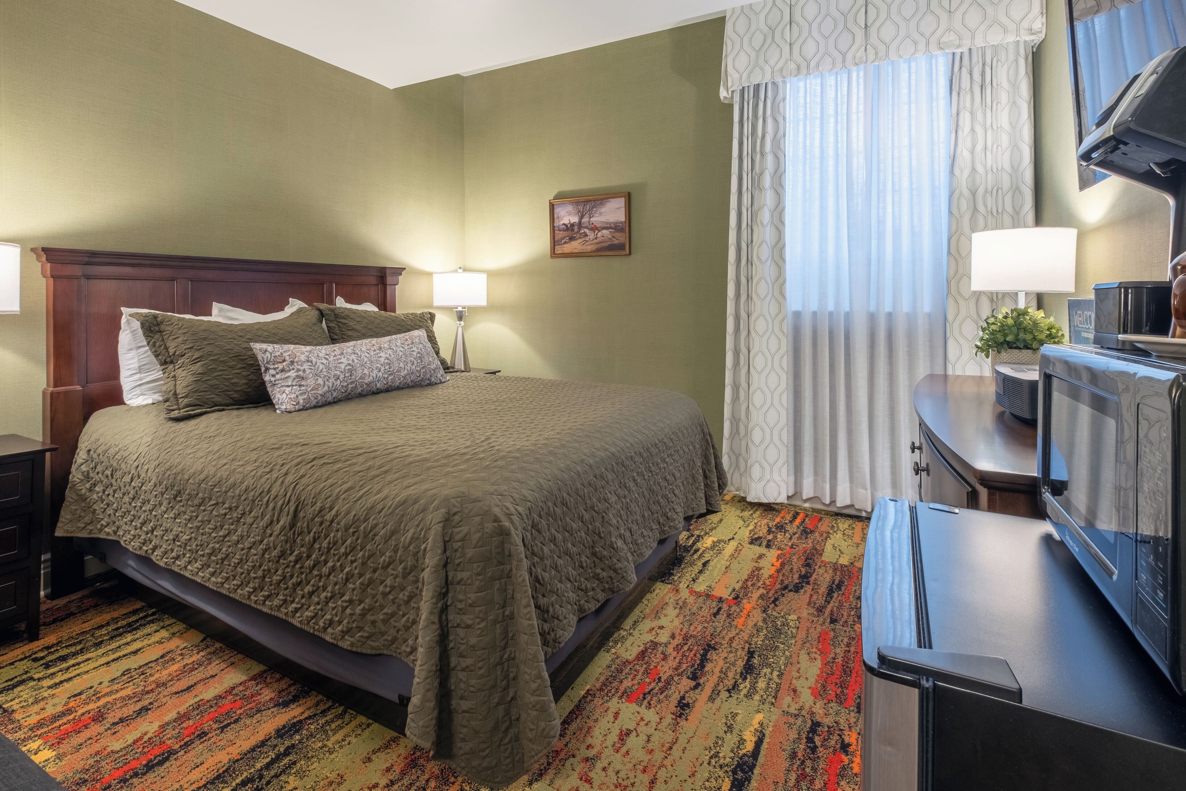 A hotel room with a bed, a desk with a television, a lamp, and a window with curtains.