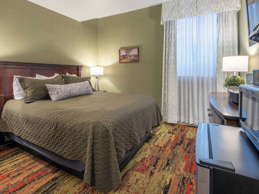 A hotel room with a bed, a desk with a television, a lamp, and a window with curtains.