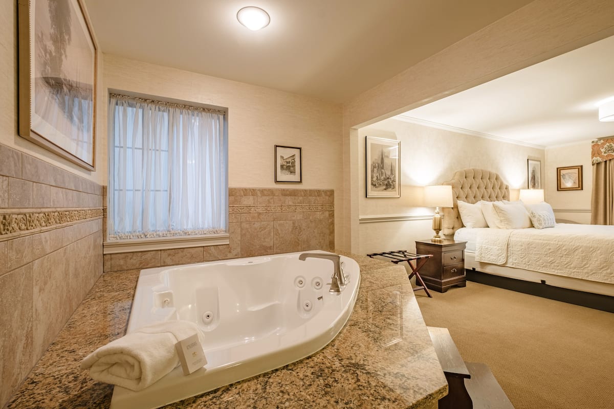 A hotel room with a large bathtub, a bed with a tufted headboard, a bedside table with a lamp, and framed pictures on the walls.