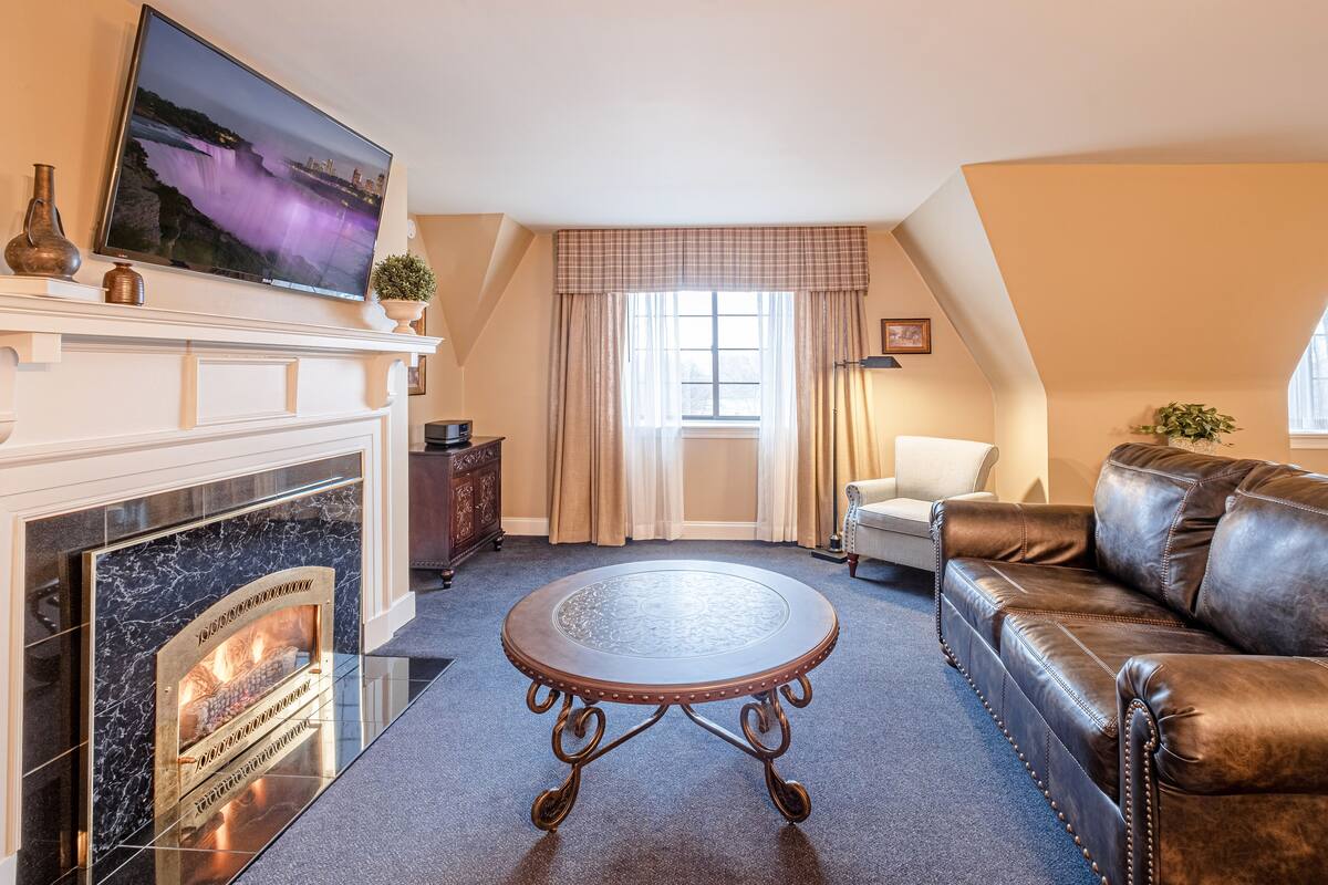 A cozy living room with a fireplace, a leather sofa, a round coffee table, and a television mounted on the wall.
