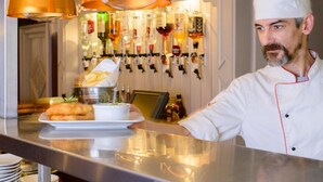 Un chef con uniforme blanco y detalles rojos prepara comida en una barra, con una variedad de platos y botellas al fondo.