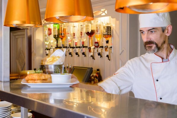 A chef in a white uniform with a red trim, preparing food at a counter with a variety of dishes and bottles in the background.
