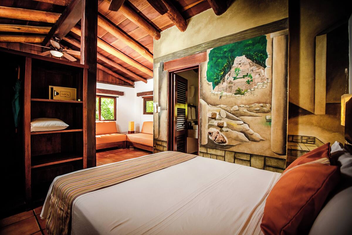 A hotel room with a bed, a wooden shelf, a mural, and a view of the outdoors.