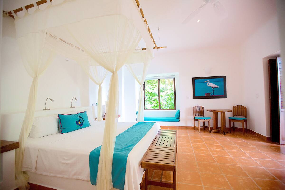 A bedroom with a bed, a bench, two chairs, a window, and a framed picture on the wall.