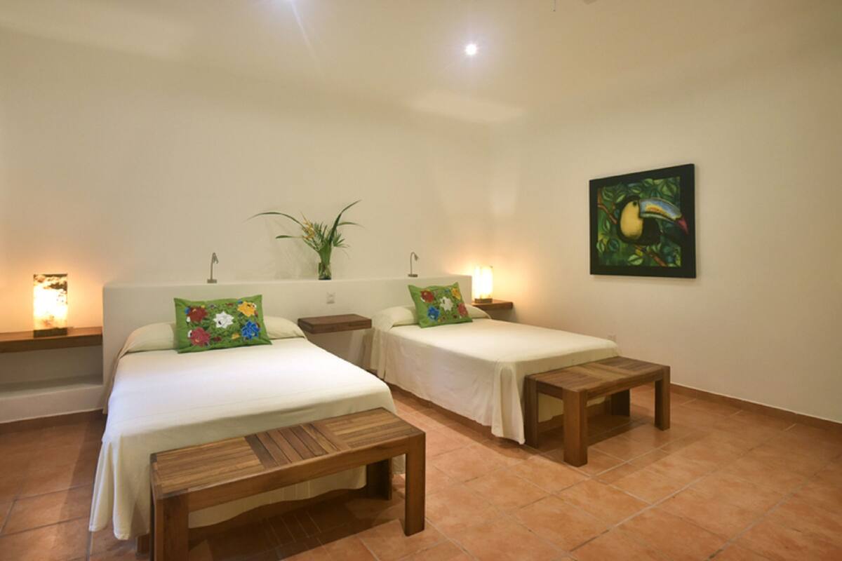 A hotel room with two single beds, a wooden bench, a small table, a plant, and a painting on the wall.