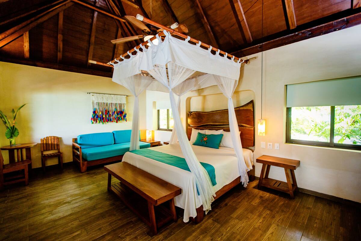 A room with a four-poster bed, wooden furniture, a bench, and a window with a view of greenery.