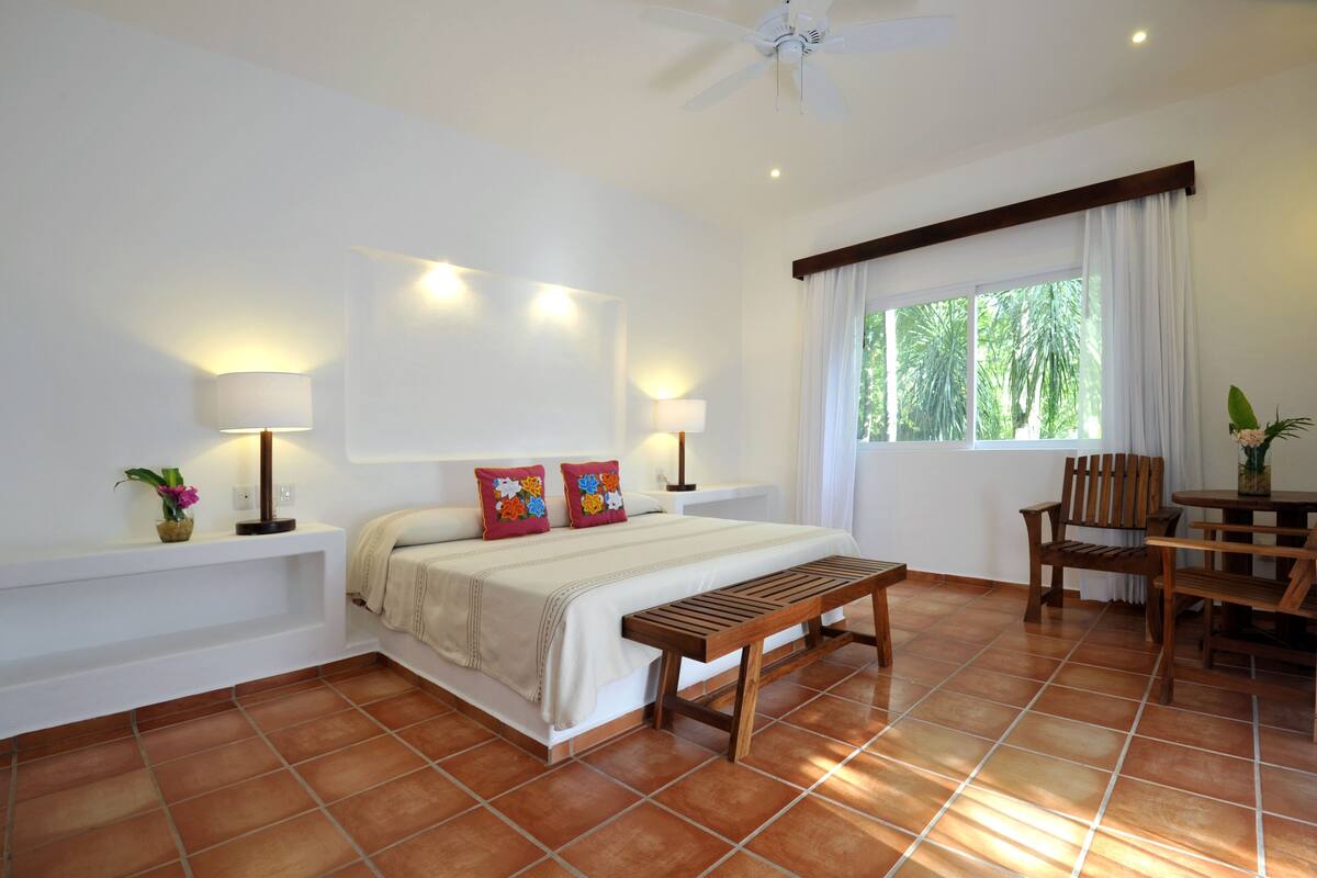 A bedroom with a bed, bedside lamps, a wooden bench, a small table, and a window with curtains.