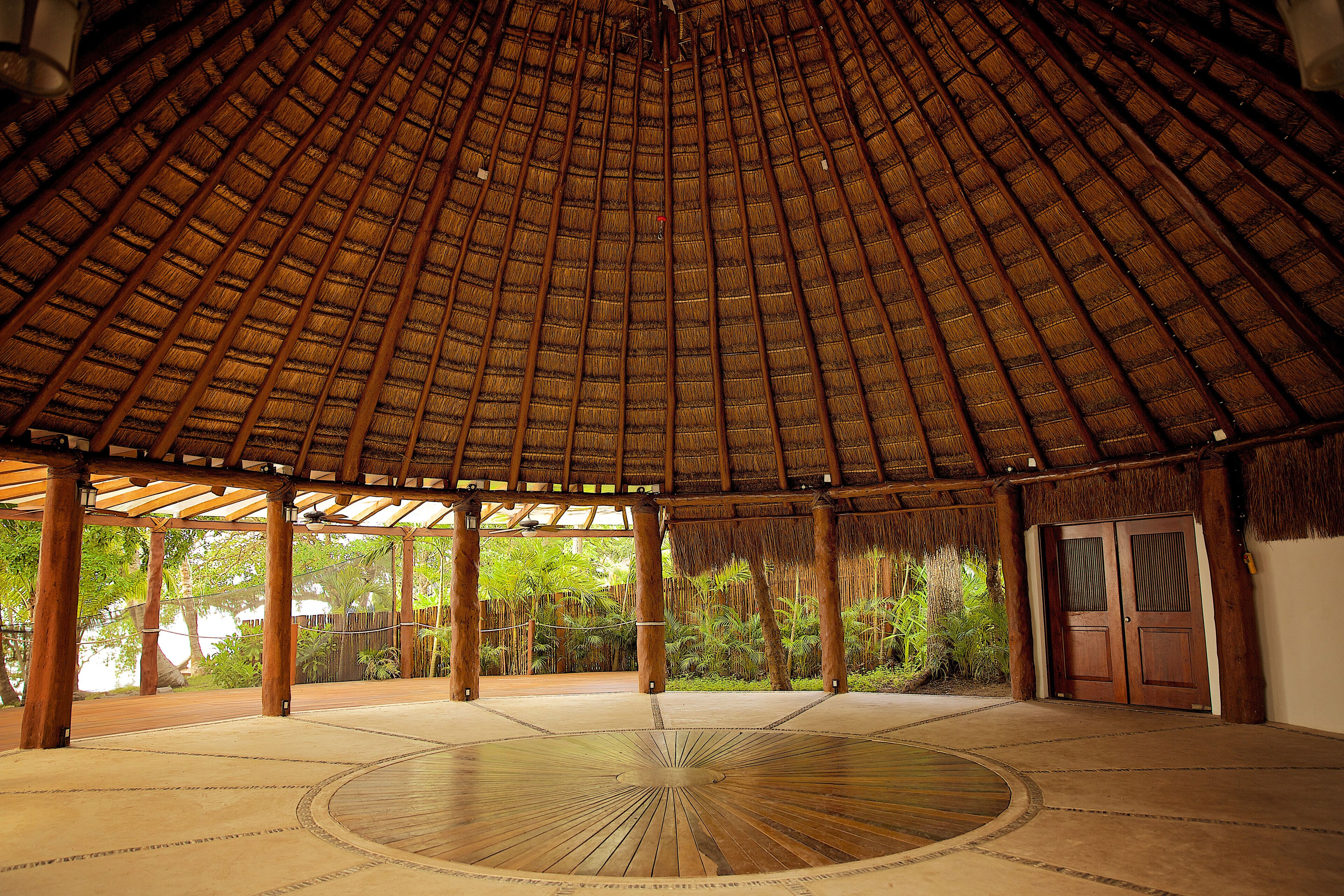 A circular wooden structure with a thatched roof, surrounded by wooden pillars and a concrete floor.