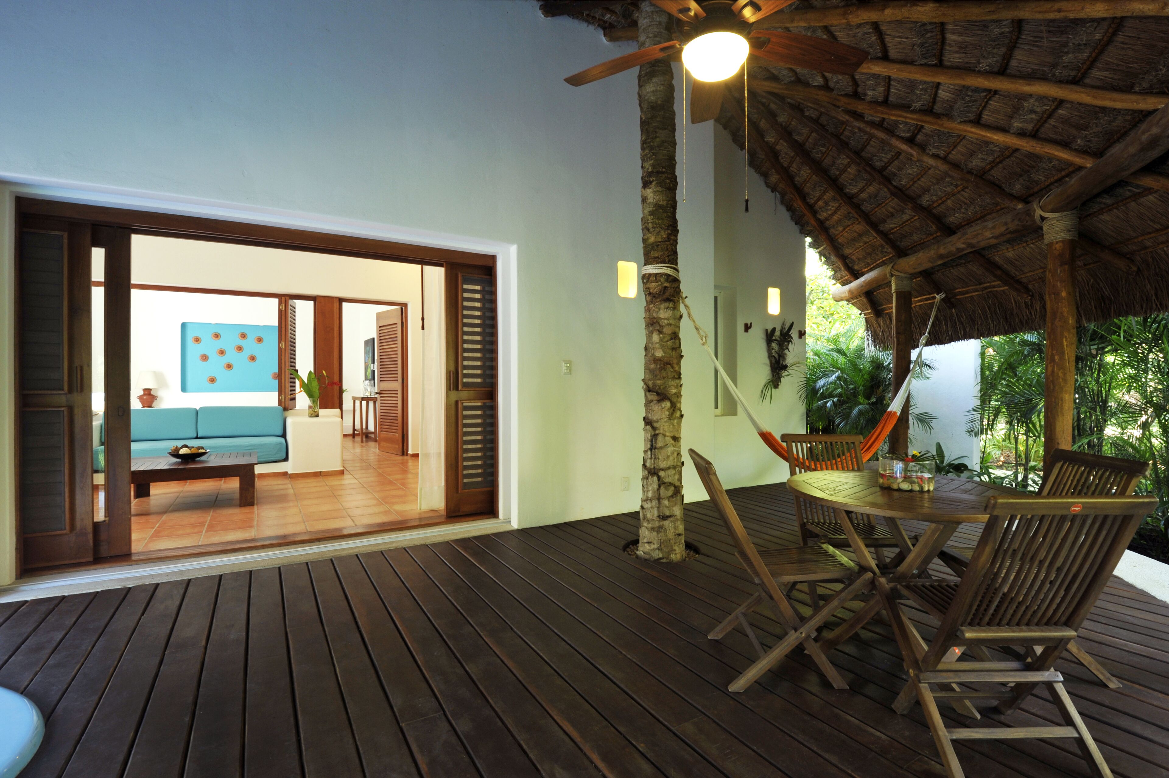 Una terraza de madera al aire libre con techo de paja, una mesa de comedor con sillas, y vista a un cuarto con sofá y pared azul.