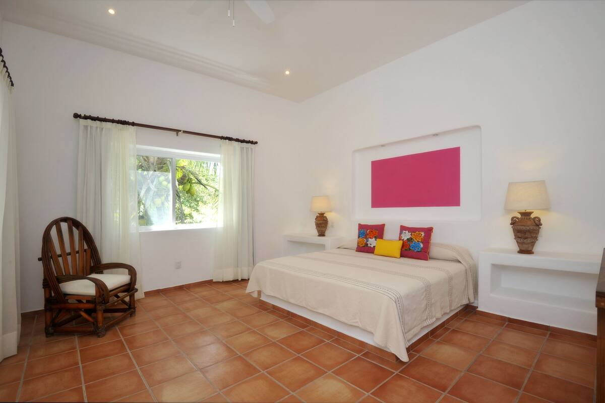 A bedroom with a bed, a chair, a window with curtains, and two bedside tables with lamps.