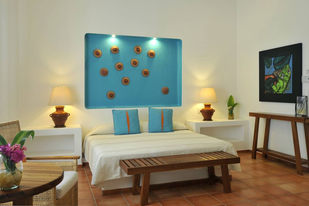 A hotel room with a bed, two bedside tables with lamps, a wooden bench, a chair, and a wall with a decorative blue panel and a framed picture.
