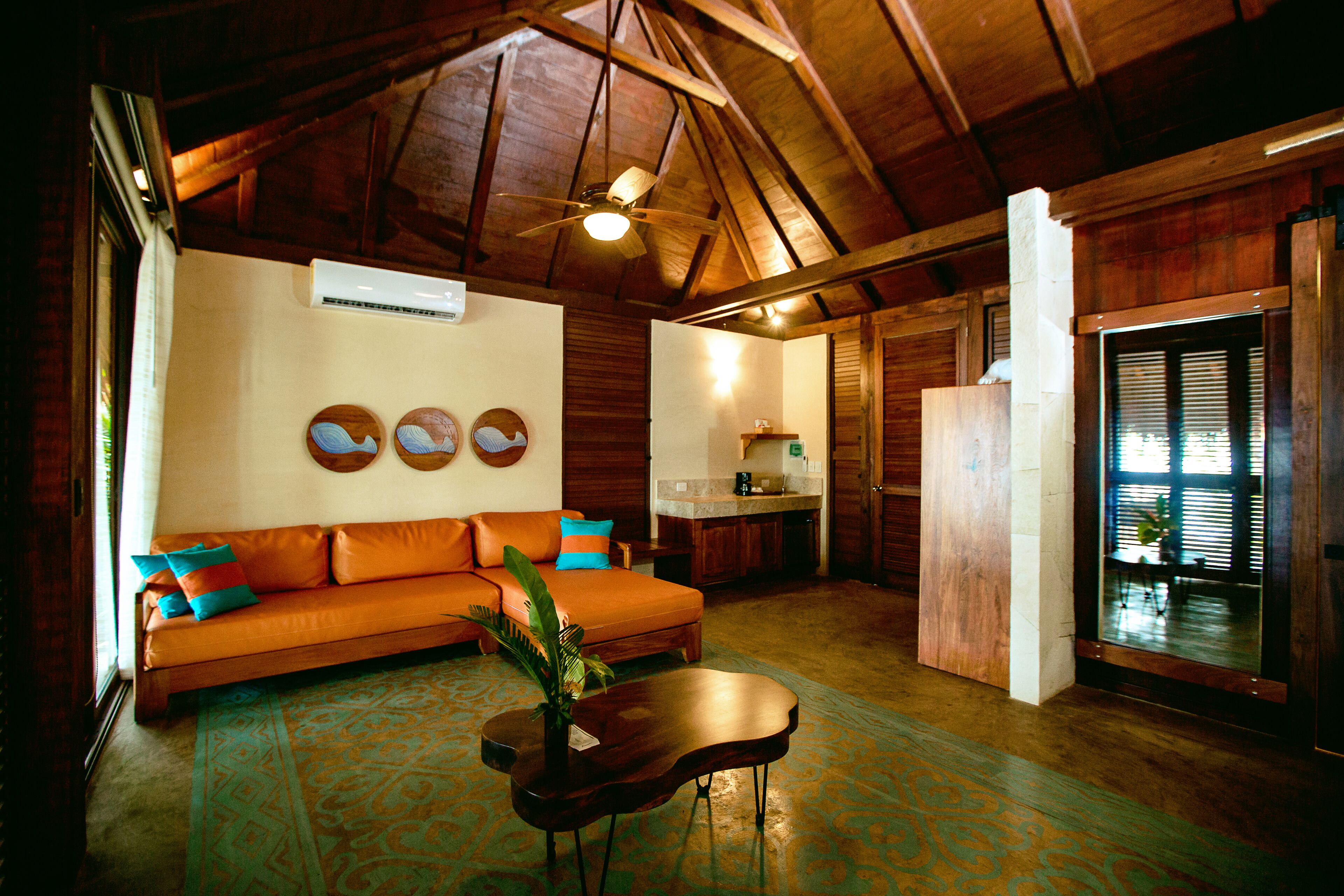 A cozy living room with a wooden ceiling, a sofa with colorful cushions, a coffee table with a plant, and a kitchen area in the background.