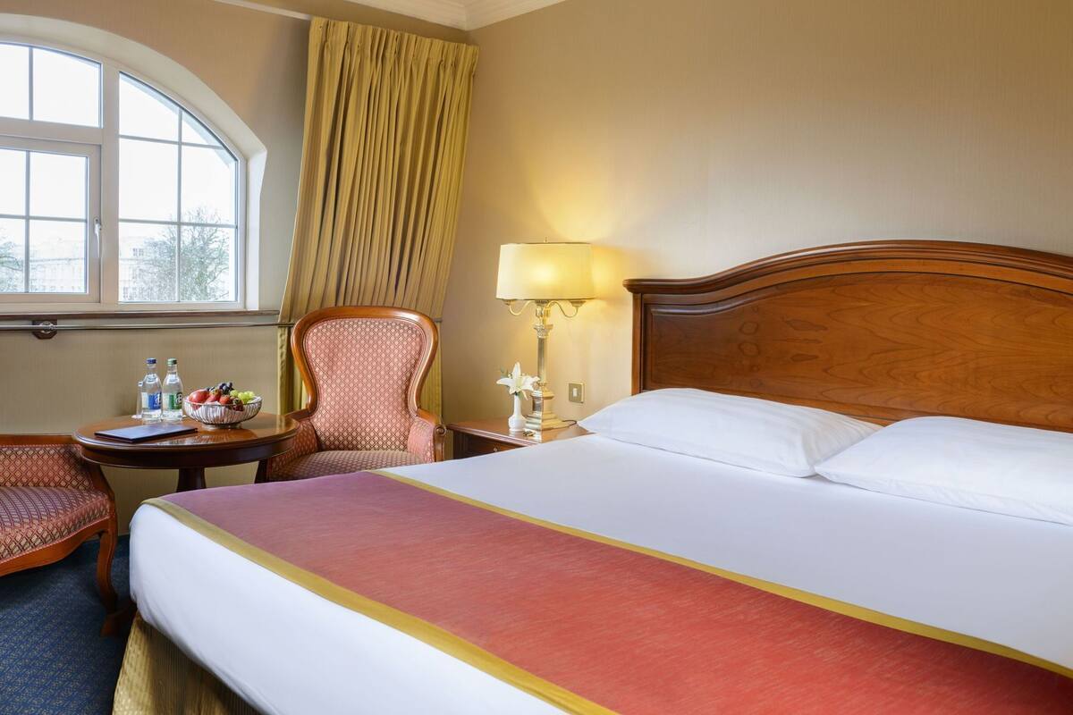A hotel room with a large bed, a desk with a lamp, a chair, and a window with curtains.