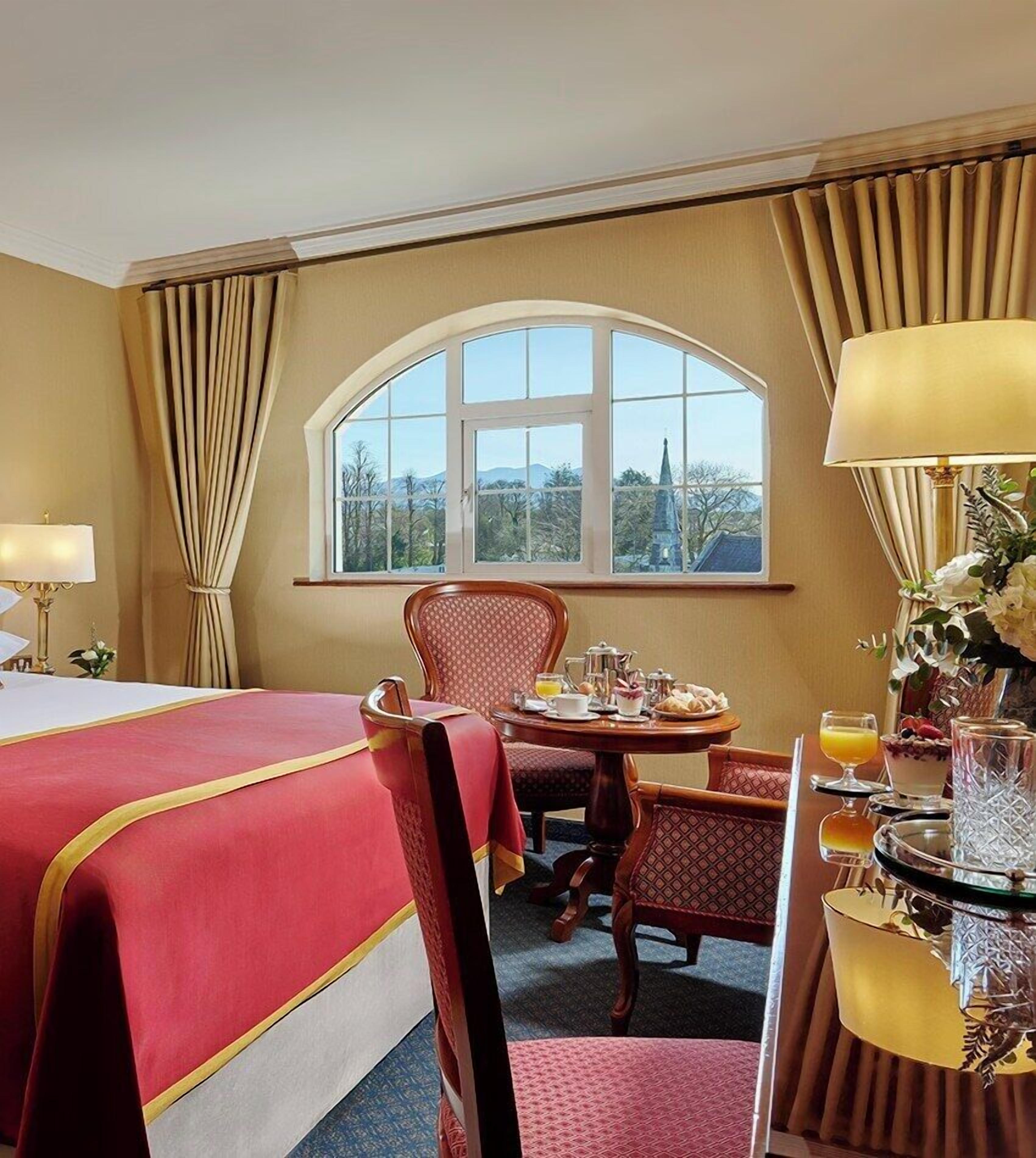 A hotel room with a large bed, a dining table with breakfast, a chair, and a view of the city through a window.