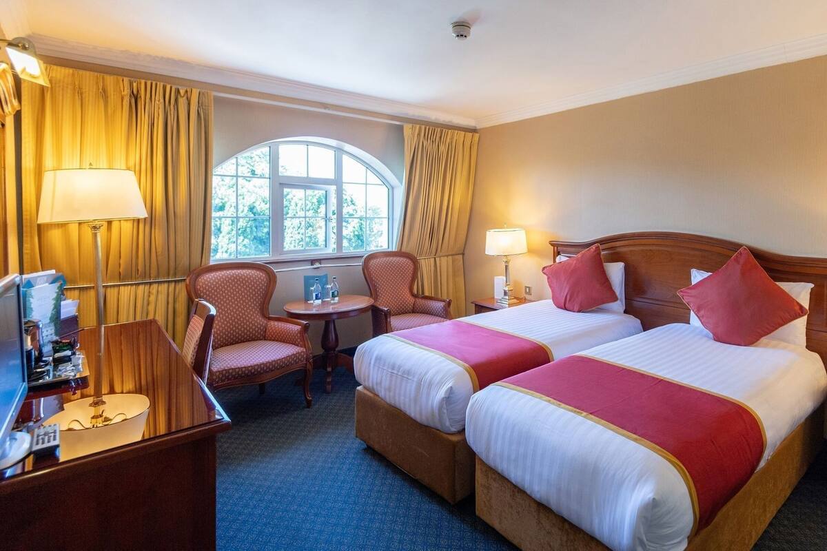 A hotel room with two beds, a desk, a chair, a window with curtains, and a lamp.