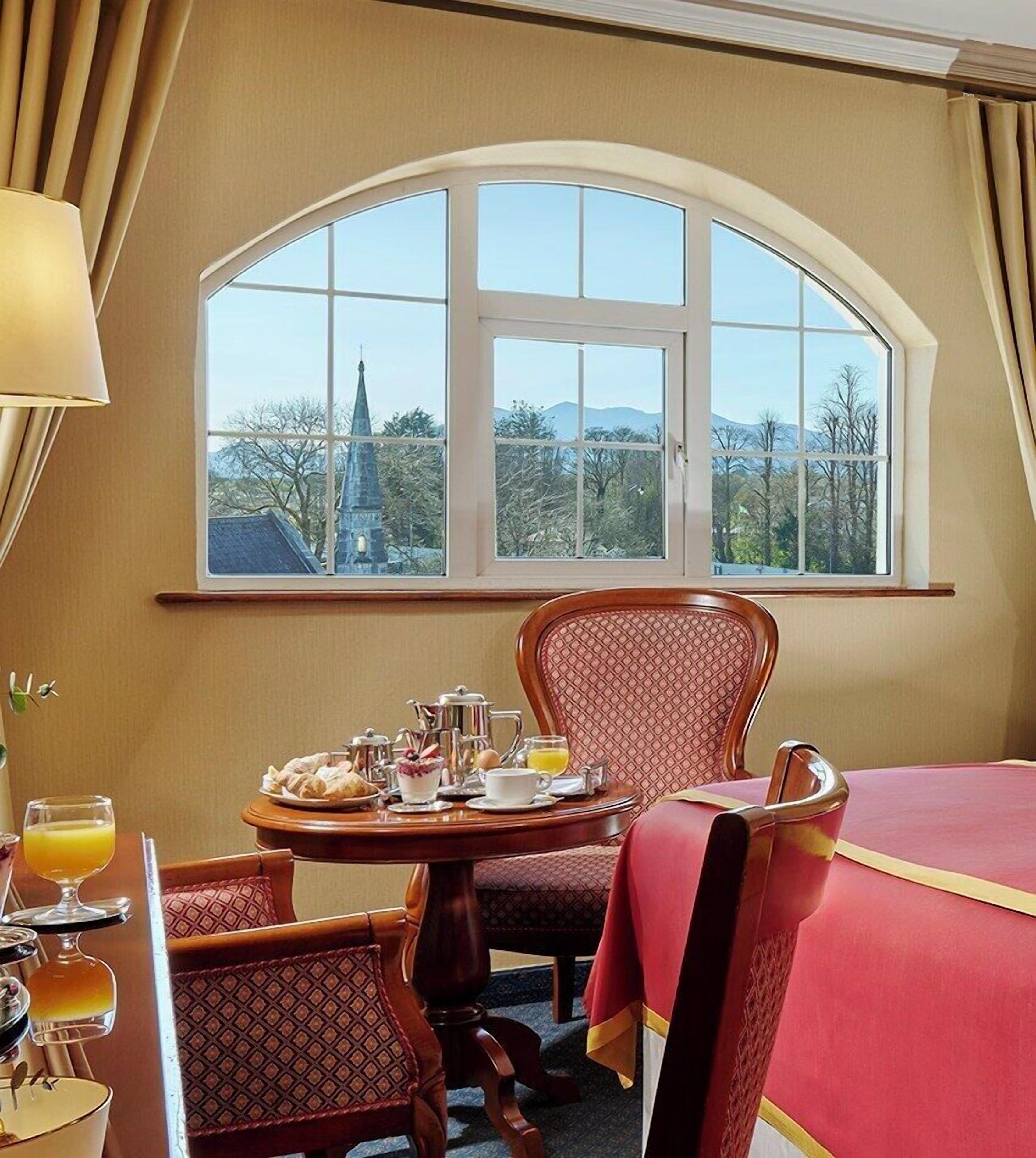 Habitación de hotel con un ventanal arqueado grande, una mesa de comedor con desayuno y una cama con colcha roja.