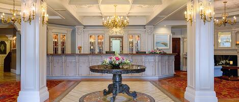 A grand lobby with a central round table, ornate chandeliers, and a reception desk.