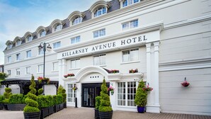 Un grand hôtel blanc à la façade classique, avec une enseigne bien visible indiquant « HOTEL KILLARNEY AVENUE » et des fenêtres disposées de manière symétrique.