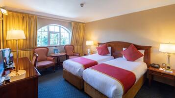 A hotel room with two beds, a desk with a lamp, a chair, and a window with curtains.
