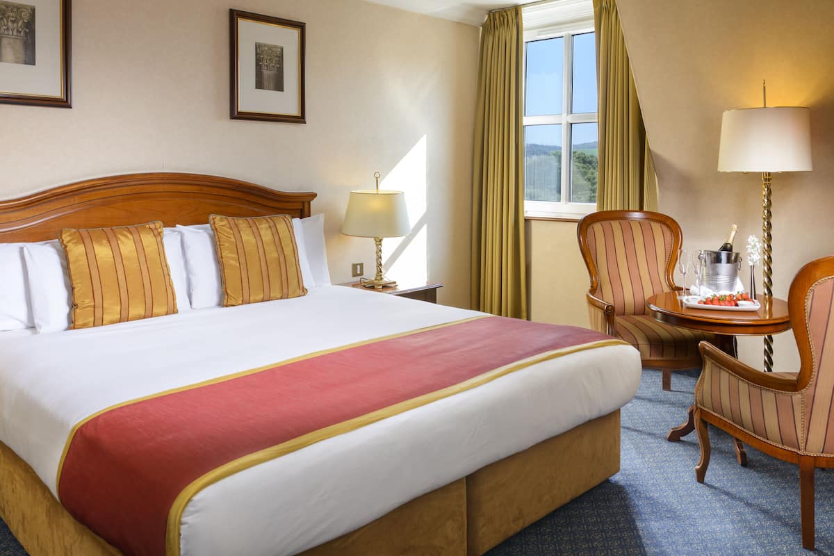 A hotel room with a large bed, a wooden headboard, a nightstand with a lamp, a chair, a table with a bottle of champagne, and a window with curtains.