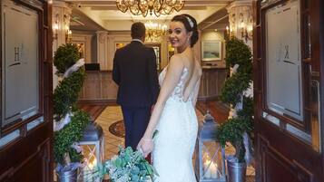A grand hallway with chandeliers, a man in a suit, and a woman in a wedding dress holding a bouquet.