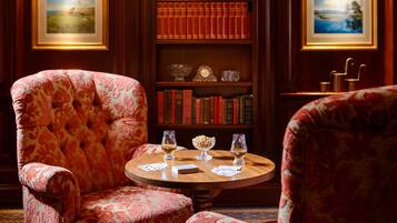 A room with a round wooden table, two armchairs, a bookshelf with books and decorative items, and framed pictures on the walls.